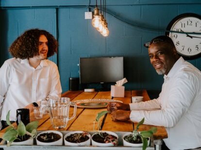 Two men sat at a table chatting and smiling to the camera