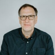 Two men sat at a table chatting and smiling to the camera